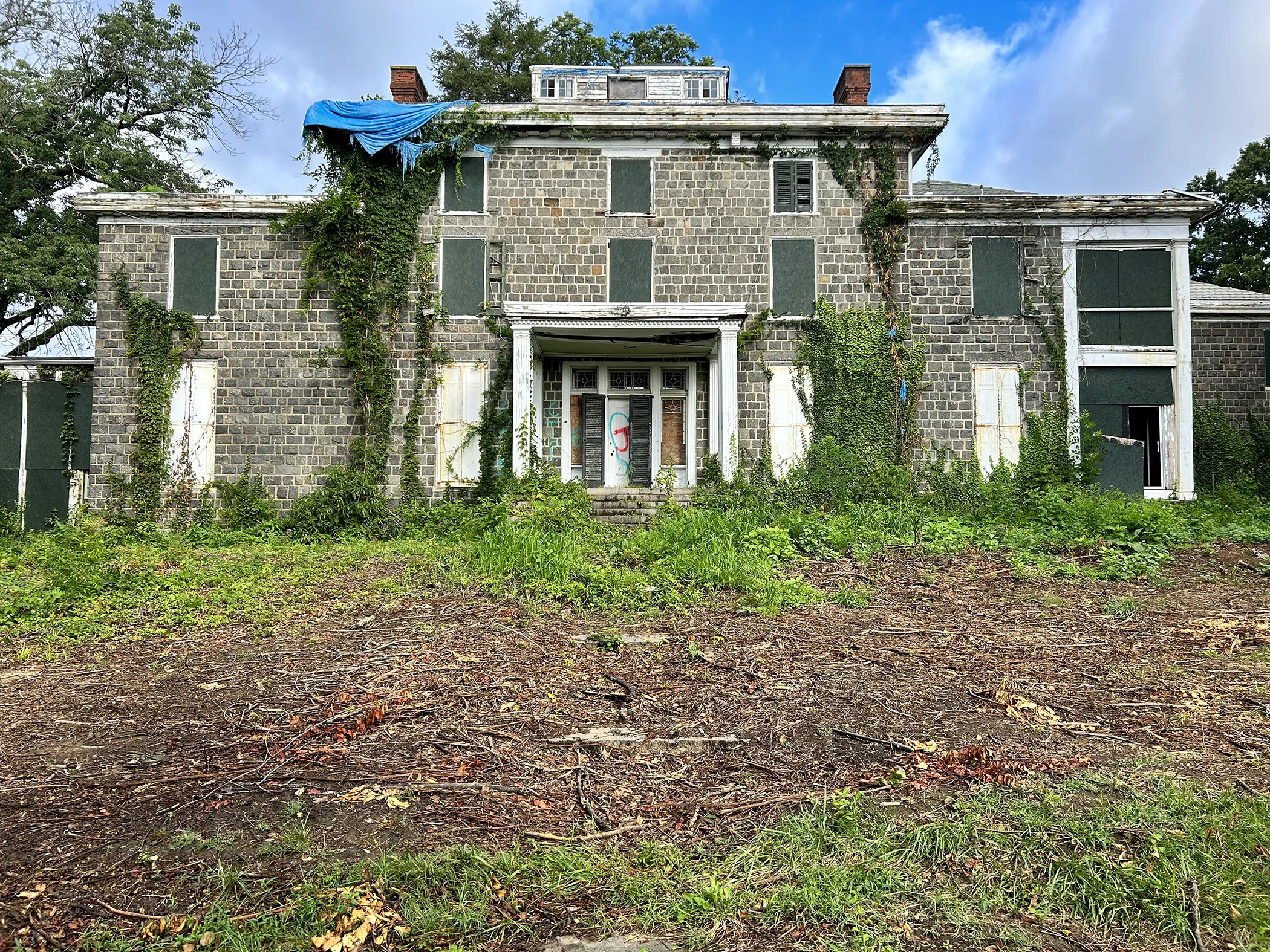 Ground-level view of Gibraltar Estate mansion facade before restoration showing vine overgrowth, boarded windows, graffiti, and abandoned condition